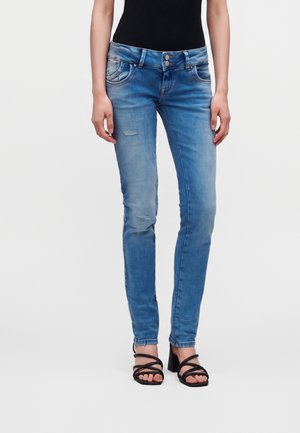 Person iført lyseblå skinny jeans med let slid, en sort ærmeløs top og sorte stropsandaler, står oprejst.