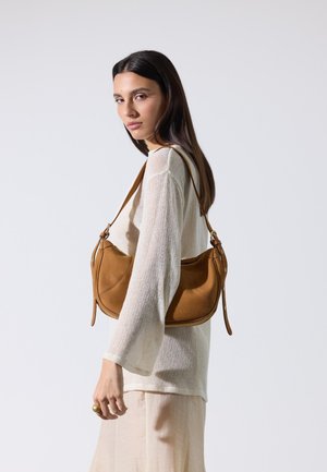 Femme aux longs cheveux foncés porte un haut beige léger à manches longues et une jupe, portant un sac à épaule en cuir marron clair sur son épaule gauche.