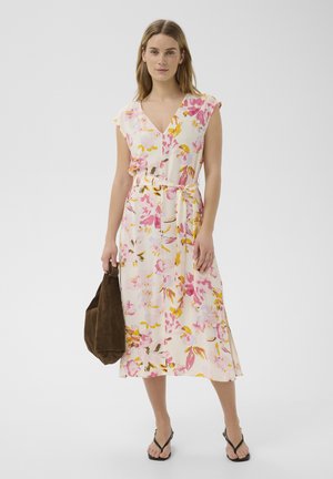 Femme portant une robe midi sans manches à motifs floraux, col en V, ceinture à la taille, tenant un sac marron et portant des sandales noires sur fond blanc.