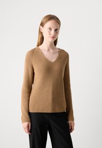 Pull en tricot col en V beige/marron, avec une texture côtelée et des manches longues, associé à un pantalon noir.