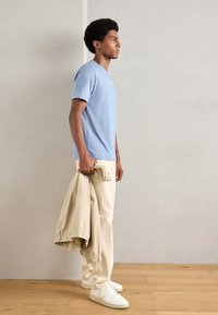 Tricou bleu deschis cu mânecă scurtă, pantaloni bej cu manșete, ținând o jachetă bej, adidași albi. Material fin cu o croială lejeră.