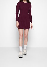 Robe bordeaux côtelée à manches longues avec une silhouette ajustée, arrivant à mi-cuisse. Portée avec des baskets de couleur claire et des chaussettes blanches.
