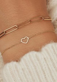 Gouden kettingarmband met een hart hanger en een schakeling in paperclip ontwerp. Draag op een pols met een zachte, lichtgekleurde gebreide trui.