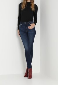 Vaqueros skinny de talle alto en azul oscuro con una textura suave, combinados con una blusa negra de manga larga de canalé y botas de tobillo rojas brillantes.