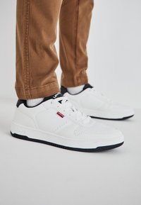Vita sneakers i syntetiskt läder med perforerade detaljer, svart sula och en kontrasterande svart hälflik; märkeslogotyp på sidan.