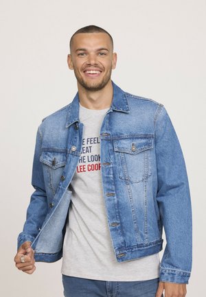 Veste en denim bleu clair avec fermeture à boutons, deux poches poitrine et coutures décoratives. Portée par-dessus un t-shirt gris à texte imprimé.