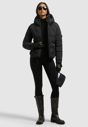 Un mannequin portant une doudoune noire, des gants, des lunettes de soleil, un legging et des bottes jusqu’aux genoux tient un petit sac noir devant un fond de studio uni.