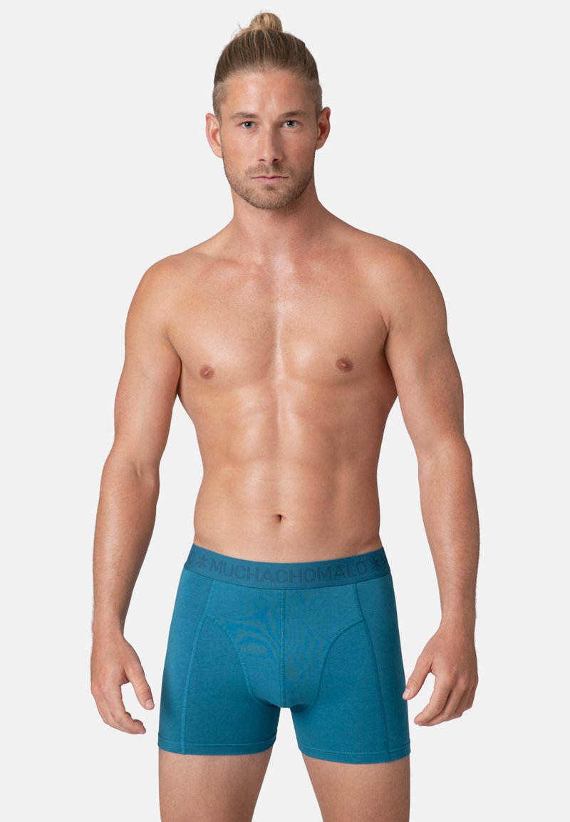 Boxer-briefs turquoise avec une taille foncée présentant le texte "MUCHACHOMALO". Fabriqués en tissu lisse et extensible avec un design ajusté.