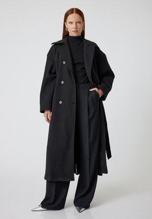 Manteau noir croisé avec larges revers, poches latérales et taille ceinturée. Porté sur un col roulé assorti et un pantalon à pattes larges, avec des chaussures pointues argentées.