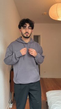 Sweatshirt cinza com capuz, bolso canguru, cordões e punhos canelados, combinado com calças pretas num chão de madeira em uma sala clara.
