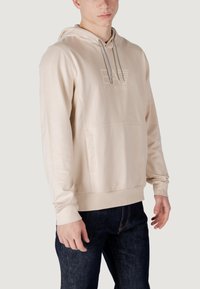 Felpa beige con cappuccio e cordino, ricamo del logo tono su tono e tasca a marsupio. Tessuto liscio e polsini a coste. Abbinata a jeans scuri.