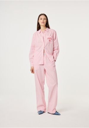 Vrouw draagt een losse, roze-wit gestreepte pyjama met een geborduurd borstzakje, staat met één hand in de zak en draagt blauwe pantoffels.