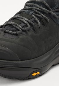 Chaussure de randonnée noire avec un upper texturé, une semelle en caoutchouc robuste et un marquage Vibram jaune vif. Les lacets sont gris avec des accents réfléchissants.
