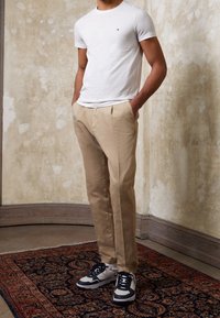 White short-sleeve t-shirt with small logo, beige pleated trousers, and black and white sneakers with textured sole, set against a neutral background.