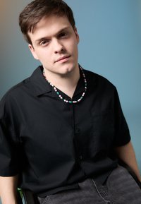 Svart kortermet skjorte med knapper, grå bukser og et flerfarget perlekjede. Bakgrunnen har en myk blå gradient.