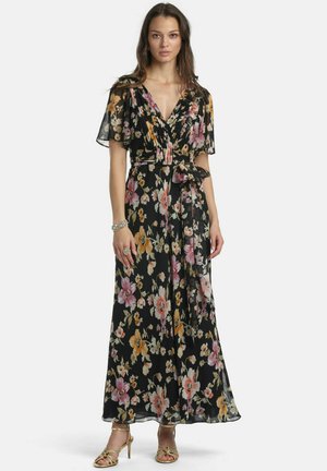 Frau trägt ein schwarzes Wickelkleid mit Blumenmuster und flatternden Ärmeln, geschnürte Taille, kombiniert mit goldenen Sandalen mit hohen Absätzen und silbernem Schmuck.