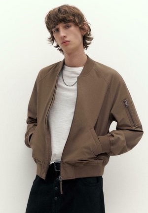 Ung person med mellemlangt brunt hår iført en brun bomberjakke over en hvid skjorte og sorte bukser, stående mod en bleg baggrund.