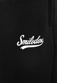 Pantalones de chándal negros con un logo blanco de "Smilodox". El material es suave con una textura lisa y el diseño incluye un ajuste estrecho.
