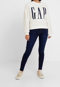 Sudadera de felpa color crema claro con la palabra "GAP" en letras marinas, combinada con jeans ajustados azul oscuro y zapatillas beige.