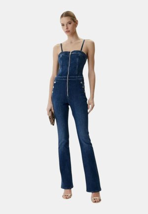 Donna che indossa una tuta in denim blu scuro con cerniera frontale, pantaloni aderenti e spalline sottili, che tiene una piccola borsa a mano marrone.