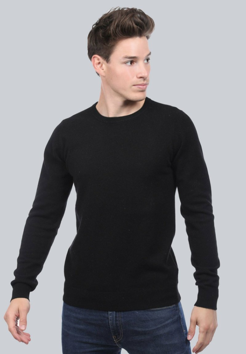 Pull en tricot noir à manches longues, col ras du cou, et texture lisse, porté sur un jean en denim bleu, contre un fond neutre.