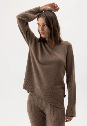 Donna che indossa un maglione marrone a coste e pantaloni abbinati, alzando un braccio sopra la testa, guardando di lato.