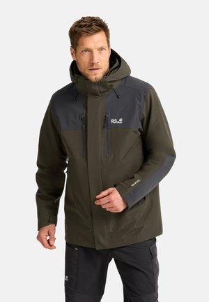 Man met een olijfgroene en donkergrijze Jack Wolfskin capuchonjas en zwarte broek, poserend tegen een effen lichte achtergrond.
