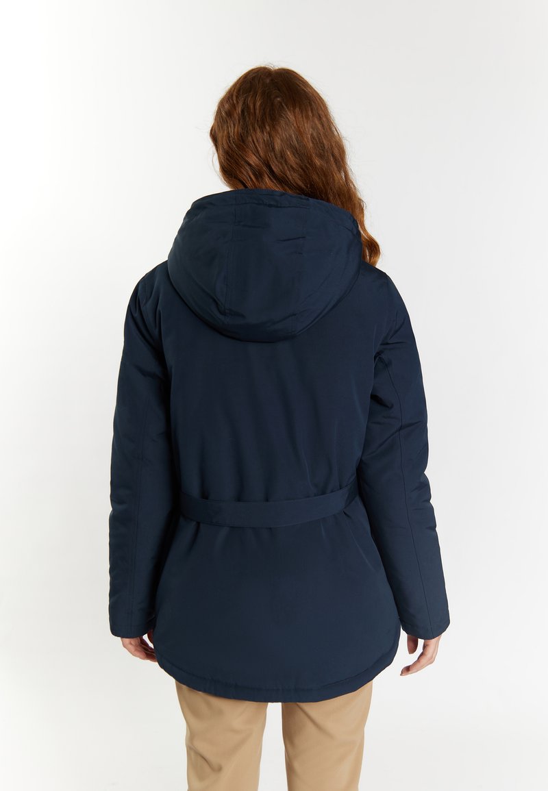 Person mit langen Haaren, der eine marineblaue Kapuzenjacke und beige Hosen trägt, von hinten betrachtet vor einem schlichten Hintergrund.