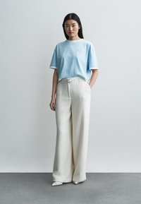 Nő áll egy egyszínű fal előtt, világoskék túlméretezett pólót, magas derekú fehér texturált bőszárú nadrágot, fehér övet és hegyes orrú cipőt visel.