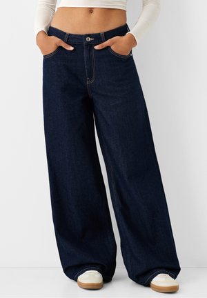 Personne portant un jean bleu foncé à jambes larges, un haut court à manches longues blanc, les mains dans les poches et des baskets blanches avec semelles en gomme.