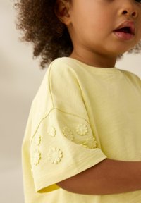 Gul bomulds T-shirt med broderede blomsterdetaljer på ærmerne, rund halsudskæring og korte ærmer. Blød, let tekstur.