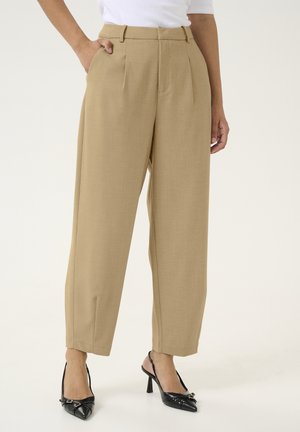 Kobieta ubrana w beżowe, plisowane spodnie z wysokim stanem i bocznymi kieszeniami oraz w czarne, spiczaste szpilki typu slingback na jednolitym tle.