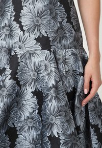 Černé šaty s velkou šedou květinovou výšivkou, s rozšířenou sukní a strukturovanou tkaninou. Detailní pohled zdůrazňuje propracované vzory květin.