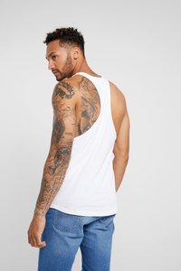 Hvid bomulds tanktop med rund halsudskæring og brede ærmegab, der viser detaljerede tatoveringer på modellens arm og skulder, parret med blå jeans.