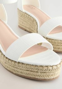 Sandalias de cuña blancas con diseño de punta abierta, hechas de un material texturizado imitación cocodrilo, con suela de yute y plantillas de un suave tono rosa.