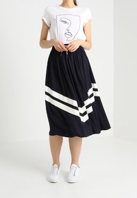 Marineblå nederdel med hvide skråstriber i kanten, parret med en hvid grafisk t-shirt. Modellen har hvide sneakers på. Løs pasform og let flydende stof.