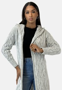 Cardigan grigio chiaro a maglia cable-knit con cappuccio, caratterizzato da un motivo testurizzato, maniche lunghe e apertura frontale, indossato sopra una maglia nera e jeans.