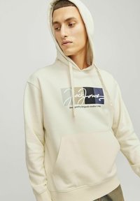 Kremowa bluza z kapturem, wykonana z miękkiego materiału, z przednią kieszenią oraz z nadrukowanym logo w czerni i kolorach. Kaptur na sznurki dla regulowanego dopasowania.