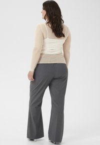 Durchsichtige beigefarbene Bluse mit subtilen Glitzereffekten, über einem figurbetonten weißen Tanktop getragen, kombiniert mit grauen Schlaghosen. Maßgeschneiderte Gestaltung, glatte Textur.