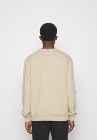 Persona con cabello corto y rizado, vistiendo un sudadera de manga larga beige y pantalones negros, frente a un fondo blanco liso.