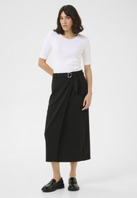 Jupe portefeuille noire avec une ceinture à boucle en D et un détail de nœud sur le côté, associée à un haut fitted à manches courtes blanc. Des mocassins noirs brillants complètent le look.