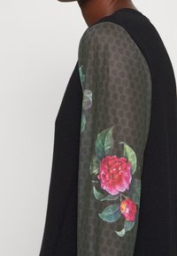 Pull noir avec une manche texturée et transparente ornée de roses roses et de feuilles sur un fond sombre à motifs.