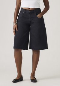 Άτομο που φορά μαύρο τζιν σορτς με φαρδιά πόδια, λευκό κοντό τοπ, μαύρες μπαλαρίνες και ασημένια βραχιόλια στον αριστερό καρπό.