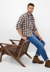 Geruite bruine en witte shirt, denim jeans, bruin suede laarzen, zittend op een houten stoel met een bruine stofzitting in een minimalistische omgeving.