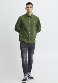 Camisa verde de botones hecha de tela texturizada, jeans grises y zapatillas negras con acentos blancos. La camisa tiene un bolsillo en el pecho y un cuello.