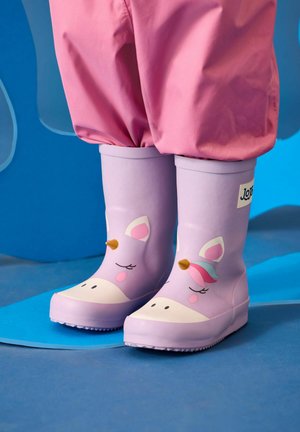 Kind draagt roze broek en lavendelroze regenlaarzen met eenhoornmotief, staand op een blauwe ondergrond met een abstracte blauwe achtergrond.