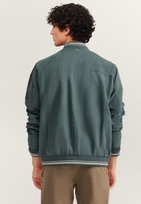 Dunkel-tealfarbige Bomberjacke mit strukturierter Oberfläche, geripptem Kragen und Bündchen sowie kontrastierenden gestreiften Akzenten. Rückansicht zeigt glatte Linien.