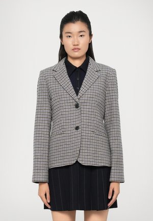 Bruin en zwart geblokte blazer met een aangeknipte kraag, twee knopen en paspelzakken, gedragen over een donker shirt en een geplooide rok.