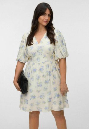 Frau mit langen dunklen Haaren, die ein leichtes, geblümtes Kleid mit Puffärmeln trägt und eine schwarze Clutch vor einfarbigem Hintergrund hält.