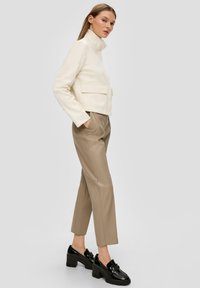 Afgesneden ivoorkleurige jas met hoge kraag en voorzakken, gecombineerd met beige faux leren broek en zwarte hakloafers.